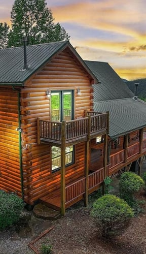 Two-story log cabin with metal roof and wraparound deck surrounded by forested mountains at sunset
