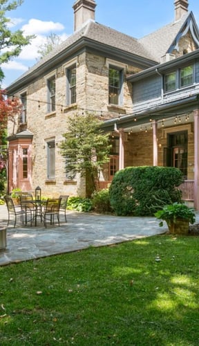 Stone Victorian house with covered porch, stone patio, outdoor seating, and manicured lawn with shrubs.