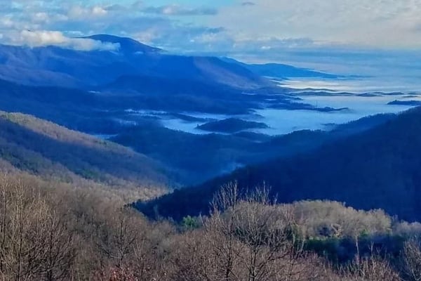 Mountain valley with bare deciduous trees, forested ridges, and distant lake visible through blue haze