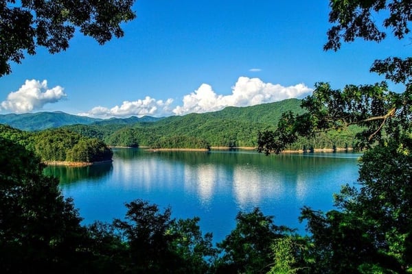Calm blue lake surrounded by forested mountains and green hills under clear sky with white clouds