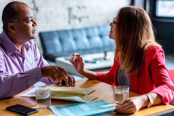 Two people in business casual attire having a discussion at a wooden table with documents and water glasses in a modern office setting.