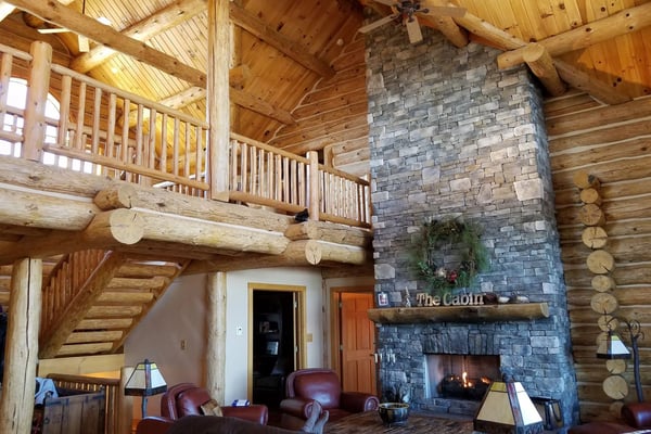 Log cabin interior with soaring timber ceiling, stone fireplace, loft with railing, and leather seating