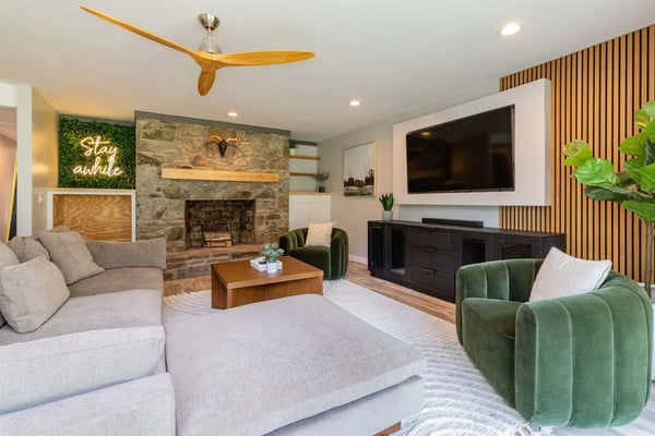 Modern living room with stone fireplace, gray and green upholstered seating, wooden ceiling fan, wall-mounted TV, and neon sign reading 'stay awhile'