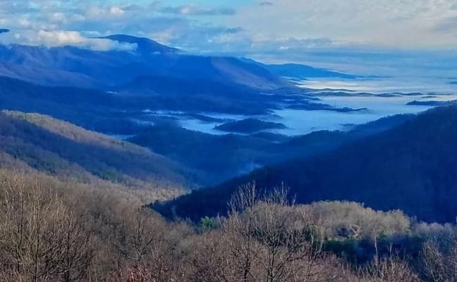 Mountain valley landscape with bare deciduous trees, forested ridges, and distant blue mountains with a lake visible in the valley below