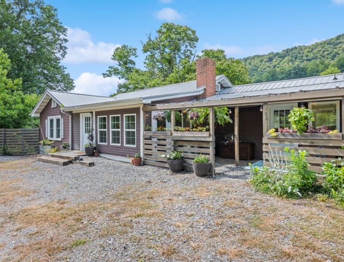 Single-story cottage with metal roof, brick chimney, and covered porch surrounded by flowering planters and forested hillside.